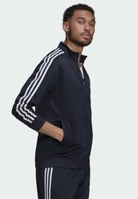 Chaqueta de chándal navy con cremallera, hecha de una tela suave, que presenta tres franjas blancas en las mangas, puños acanalados y bolsillos laterales.