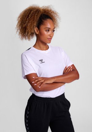 White cotton t-shirt with short sleeves, a round neckline, and side black accents. Features a small logo on the chest. Worn with black joggers.