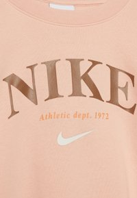 Růžová mikina se velkým hnědým nápisem "NIKE", menším oranžovým textem "Athletic dept. 1972" a bílým logem swoosh Nike. Měkká textilie.