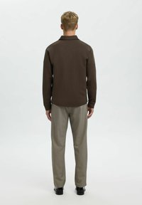 Camicia marrone a maniche lunghe con colletto, abbinata a pantaloni grigi chiari. L'outfit presenta una vestibilità rilassata e una texture morbida.