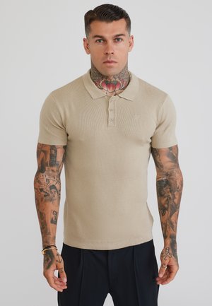 Uomo con tatuaggi sul collo e sulle braccia, che indossa una polo beige a maniche corte e pantaloni neri, in piedi davanti a uno sfondo chiaro e uniforme.