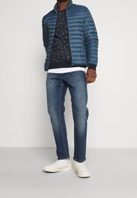 Blue puffer jacket with horizontal quilting, a zip front, and stand collar, layered over a dark patterned sweater, paired with blue jeans and white sneakers.