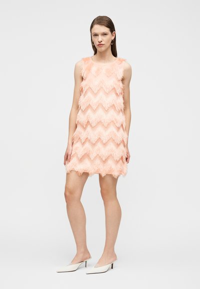 Γυναίκα που φοράει ένα μίνι φόρεμα χρώματος ροδάκινου χωρίς μανίκια με κρόσσια και λευκές μυτερές γόβες με χαμηλό τακούνι, στέκεται μπροστά σε ένα απλό φόντο.