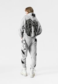 Sudadera gris con bordado de alas de ángel negras en la espalda, combinada con pantalones a juego que presentan diseños desgastados de alas y correas de utilidad.