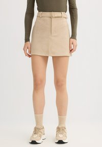 Minifalda de pana beige con diseño ajustado, cintura con cinturón y dos bolsillos delanteros. Se lleva con zapatillas beige y calcetines claros.