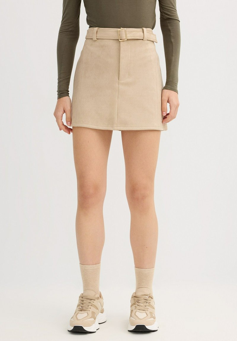 Minifalda de pana beige con diseño ajustado, cintura con cinturón y dos bolsillos delanteros. Se lleva con zapatillas beige y calcetines claros.