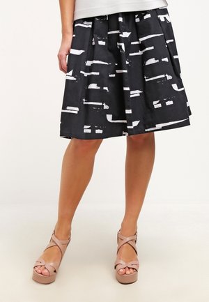 Jupe plissée noire avec des motifs abstraits blancs, fabriquée en tissu léger ; associée à des sandales à plateforme nudes avec des brides croisées.
