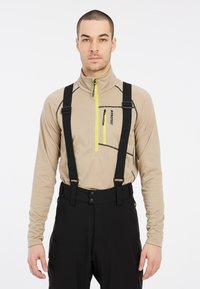 Camiseta beige de manga larga con cuello de media cremallera, con detalles en negro; combinada con pantalones negros asegurados con correas y clips.