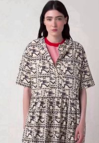 Korte mouwen jurk van licht beige stof met een geometrisch patroon in donkerbruin en blauw, met een knoopsluiting aan de voorkant en een geplooide taille.