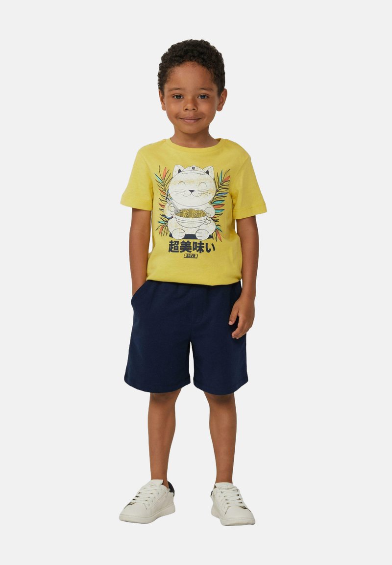 T-shirt di cotone giallo con una grafica di un gatto con noodles, abbinato a pantaloni corti blu navy. Il modello indossa sneakers bianche. Design semplice e casual.