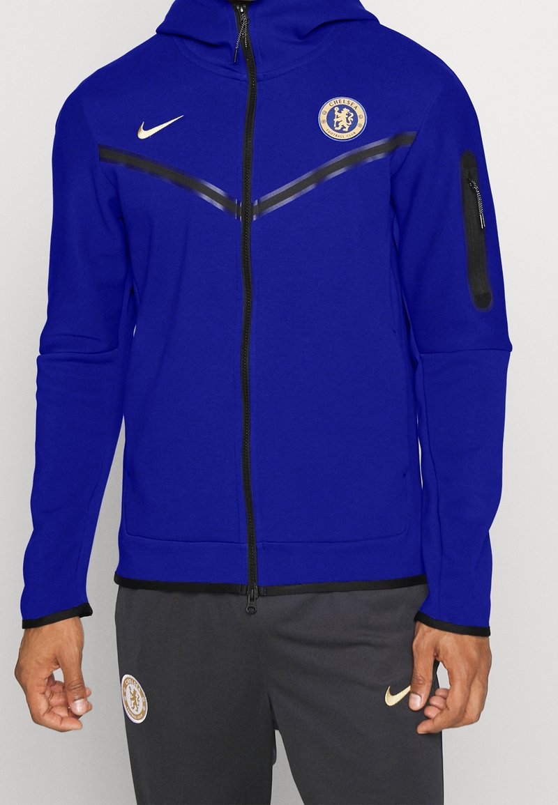 Homme portant un sweat à capuche bleu zippé avec les logos de Chelsea FC et Nike, associé à un pantalon gris foncé arborant les mêmes logos.