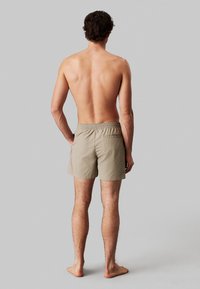Pantaloncini da bagno beige con vita elastica, tasca posteriore e gamba corta, indossati da un modello maschile visto dalla schiena.