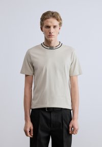 Μπεζ κοντομάνικη μπλούζα από απαλό ύφασμα, με μαύρο ριμπ γιακά και την επωνυμία "BOSS" στο λαιμό, συνδυασμένη με μαύρο παντελόνι.