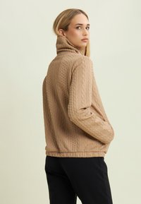 Jeune femme portant un pull col roulé en maille torsadée marron clair et un pantalon noir, se tenant avec les mains dans les poches, regardant par-dessus son épaule.