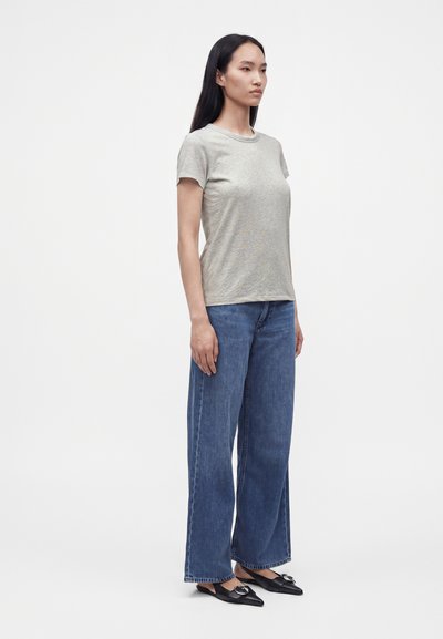 Femme debout portant un t-shirt gris clair à manches courtes, un jean bleu à jambes larges et des ballerines noires pointues avec un détail de boucle.