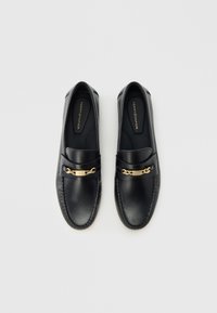 Μαύρα δερμάτινα μοκασίνια με χρυσή διακοσμητική αλυσίδα, με ραφές κατά μήκος των ακρών και λεία υφή. Μαλακά εσωτερικά πέλματα με ορατή μάρκα.