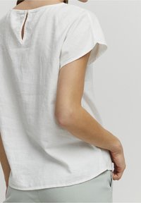 Weißes Leinen-Top mit kurzen Ärmeln, rundem Ausschnitt und einem Schlitzdetail am Rücken. Leicht geschwungener Saum. Glatte Textur.