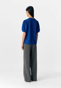 Blauwe getextureerde blouse met poffende mouwen, gecombineerd met grijze wijde broekspijpen. De blouse heeft een patroon en een ronde halslijn.