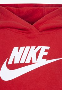 Sarkans hoodijs, kas izgatavots no mīkstas auduma, ar lielu baltu Nike logo vidū krūtīm. Tam ir kenguru kabata un kapuce ar auklu.