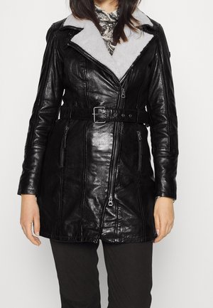 Woman wearing a black leather belted jacket with a white shearling collar over a patterned top, paired with black pants.
