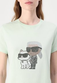 Lys myntgrønn t-skjorte med grafikktrykk som viser en katt og en kledd figur i solbriller, utsmykket med svarte krystaller.