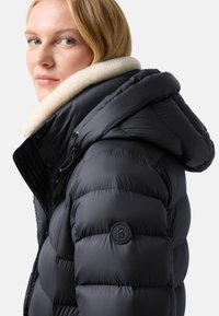 Černá péřová bunda s bílým umělým kožešinovým límcem, se zipovým uzávěrem, bočními kapsami a kulatým logem na rukávu.