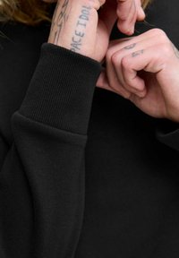 Puño de sudadera negra con textura acanalada, con tatuajes expuestos en una mano cercana. Enfoque en la tela y los detalles.