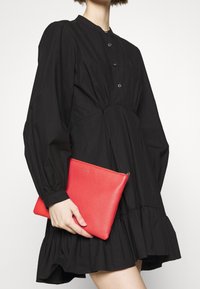 Sort kjole med pufærmer og knapdetaljer, parret med en rød læderclutch med en glat tekstur og lynlåslukning.