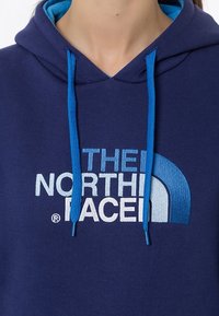 Granatowy hoodie z jasnoniebieskimi akcentami i sznurkami. Posiada logo "THE NORTH FACE" w białym kolorze oraz zakrzywiony wzór graficzny z przodu.