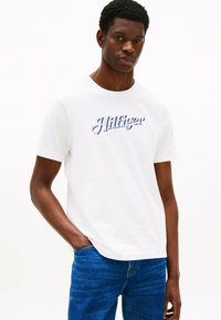Hvid bomulds t-shirt med et blåt broderet "Hilfiger" logo på brystet. Den har en rund halsudskæring og korte ærmer.