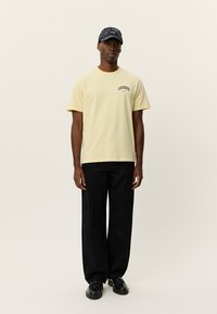 Κίτρινη βαμβακερή μπλούζα με την επιγραφή "LES DEUX" στο στήθος, συνδυασμένη με μαύρο παντελόνι με σχέδια και μαύρα παπούτσια, φορεμένη με σκούφο σε σκούρο χρώμα.