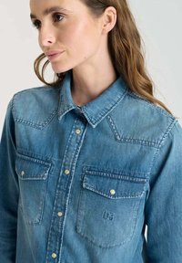 Kvinna iklädd en ljusblå denimskjorta med framknappar och två bröstfickor med luckor, som vänder sig något åt vänster.