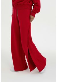 Rote weite Hosen aus weichem Stoff mit einem Reißverschluss vorne und seitlichen Schlitzen. Tragen Sie weiße Sneaker, die das legere Design unterstreichen.