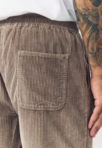 Braune Cordhosen mit gerippter Textur, einer Gesäßtasche und einem elastischen Bund, teilweise sichtbar mit einer Hand auf der Hüfte.