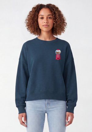 Femme aux cheveux bouclés portant un sweat-shirt bleu foncé avec un petit motif coloré de distributeur de boules de gomme sur la poitrine gauche et un jean bleu clair.