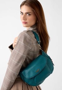 Torba na ramię z turkusowej skóry o fakturowanej powierzchni, z klapką zapinaną na zatrzask oraz złotym logo, zawieszona na kontrastowym pasku.