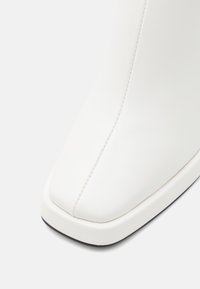 Bottine blanche à cheville avec une texture lisse rappelant le cuir, un bout carré, des détails de couture subtils et une semelle noire contrastante.