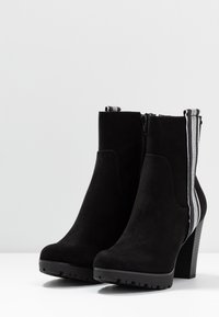 Botines de ante negro con tacón grueso, que cuentan con una cremallera lateral y un detalle de franja contrastante a lo largo de la parte posterior. Suela texturizada para agarre.