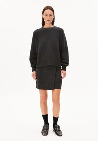Frau in einem dunkelgrauen oversized Pullover und einem passenden Rock, mit schwarzen Socken und genagelten schwarzen Slippern, die vor einem weißen Hintergrund steht.