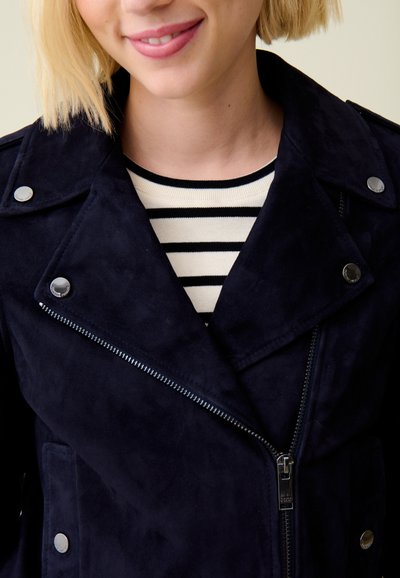 Veste en daim bleu marine avec quincaillerie en argent, fermeture éclair à l'avant et boutons-pression. Portée par-dessus une chemise à rayures noires et blanches.