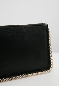Pochette noire texturée avec bordure en chaîne dorée et logo "Forever New" discrètement embossé, sur un fond uni.