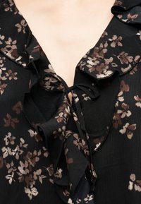 Μαύρη μπλούζα με καφέ και μπεζ λουλουδένια σχέδια, με λαιμόκοψη με βολάν που δένει στο μπροστινό μέρος με λεπτή υφασμάτινη κορδέλα.