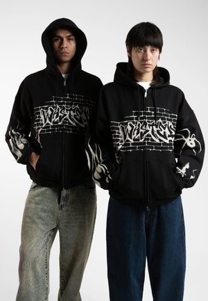 Sorte zip-up hettegensere med graffiti-stil design i hvitt, med et murveggmønster og edderkoppdetaljer på ermene, båret over løse jeans.