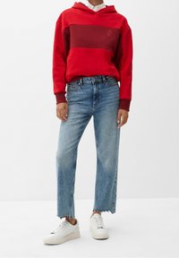 Sudadera con capucha roja y burdeos con un logotipo, combinada con jeans de tiro alto de color azul claro con dobladillos deshilachados y zapatillas blancas.