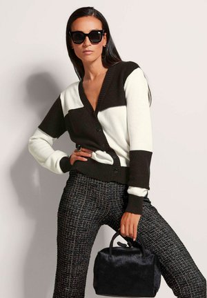 Schwarz-weiße Strickjacke mit V-Ausschnitt und Knopfverschluss, kombiniert mit strukturierten schwarzen Hosen und einer kleinen schwarzen Pelztasche.