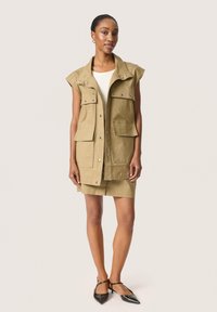 Khaki mouwloze utility vest met vier voorzakken, zilveren drukknoopsluitingen en een gestructureerde kraag, gecombineerd met een bijpassende rok en zwarte schoenen.