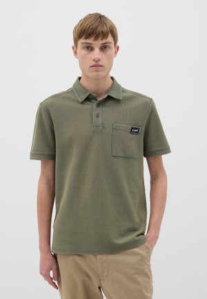 Polo de color verde oliva hecho de una tela texturizada, con un cierre de tres botones, mangas cortas y un solo bolsillo en el pecho con un parche de logo.