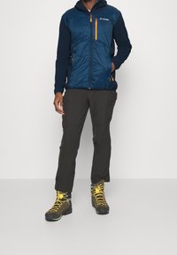 Man som bär en marinblå jacka med orange dragkedjor, svarta byxor och bruna vandringskängor med gula snören, stående mot en vit bakgrund.