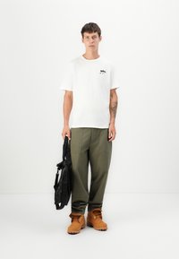 White short-sleeve t-shirt with a small black graphic on the chest, olive green loose-fitting pants, and brown lace-up boots. Black shoulder bag.