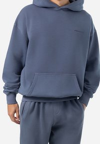 Sudadera con capucha en material gris sólido, con un bolsillo frontal, puños acanalados y un detalle sutil de logo cerca del cuello.